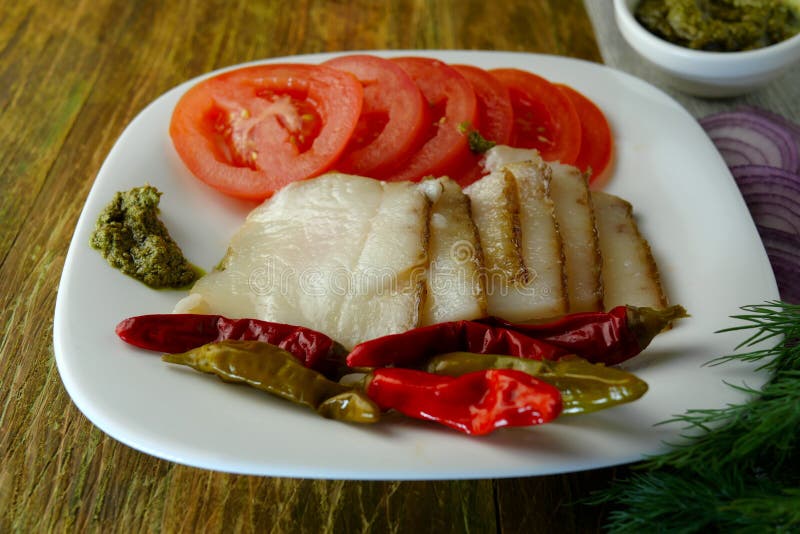 Appetizing snack. Sliced ​​smoked pork lard, tomatoes, pickled chili peppers and pesto sauce lie on brown wooden surface stock photo