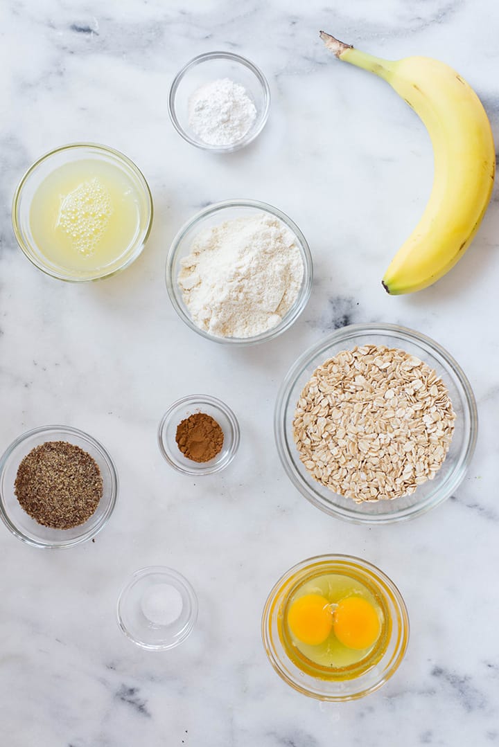 An overhead image of a kitchen counter with all the ingredients for the Banana Protein Pancakes including uncooked rolled oats, a ripe banana, eggs, egg whites, baking powder, sea salt, ground cinnamon, vanilla or unflavored protein powder and ground flaxseeds.