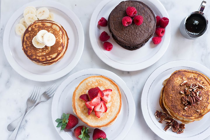 An overhead image of a kitchen counter with 4 different kinds of Protein Pancakes, including Banana Protein Pancakes, Chocolate Protein Pancakes, Sweet Potato Protein Pancakes and Oatmeal Protein Pancakes.