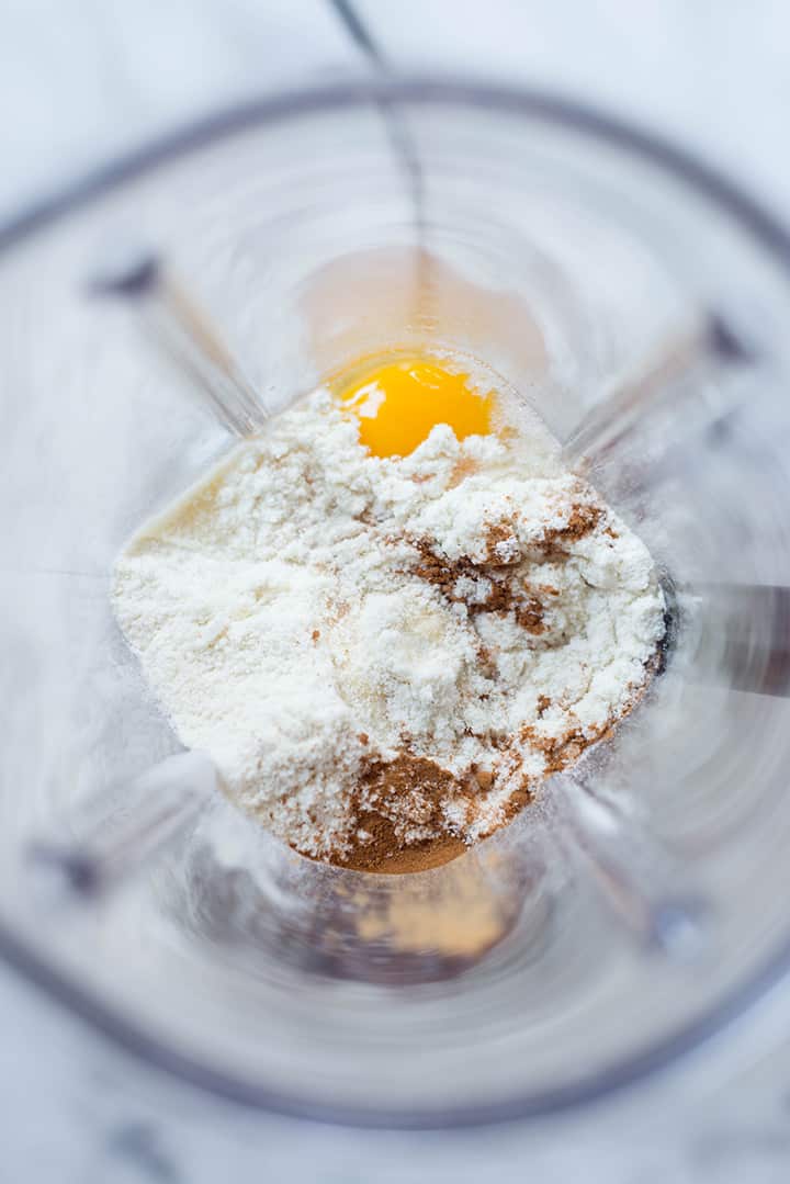 An overhead image of a blender with all the ingredients for the Oatmeal Protein Pancakes ready to be blended, including eggs, uncooked rolled oats, protein powder, pure maple syrup and ground cinnamon.